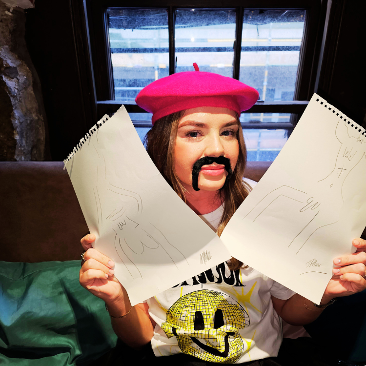Female guest in a red beret and fake moustache proudly displaying her artwork at a Hen Party Life Drawing in Hersham. Hen Party Life Drawing in Hersham near me