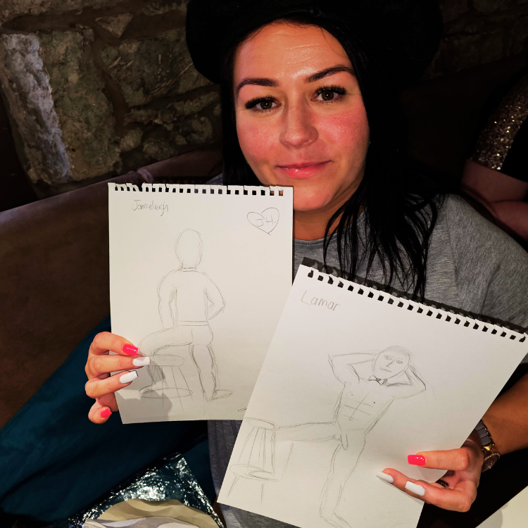 A guest in a black beret holding up two drawings at a hen party life drawing in Lyndhurst. Hen party life drawing class near me.
